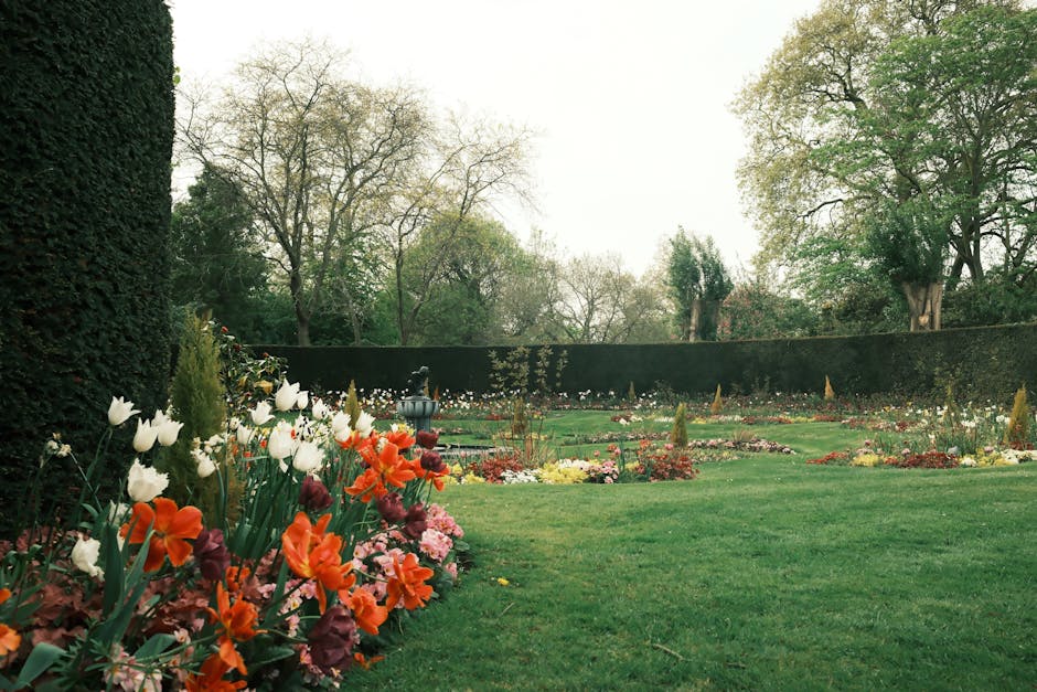 A historic garden featuring a lush, well-maintained lawn bordered by a variety of flowering plants and shrubs. In the foreground, a stone pedestal supports a decorative birdbath, surrounded by purple